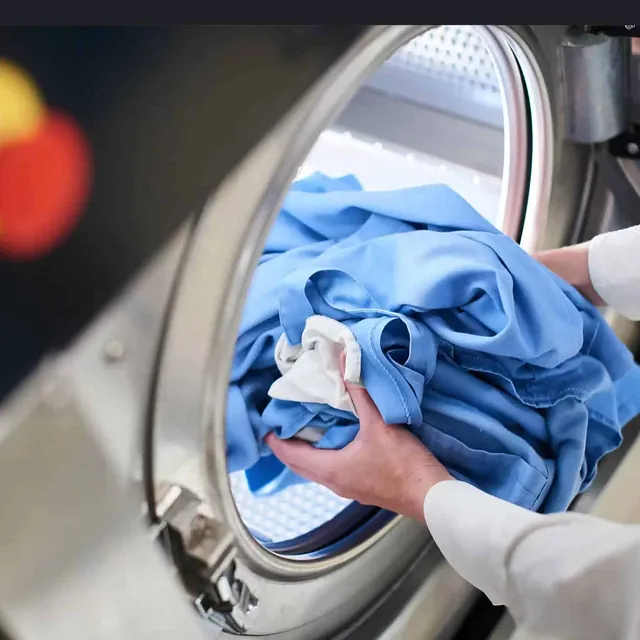 En person legger blå arbeidsklær i en industriell vaskemaskin.